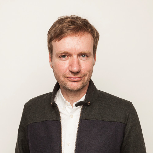 Eine Person mit braunen Haaren in einem Jackett vor neutralem Hintergrund.