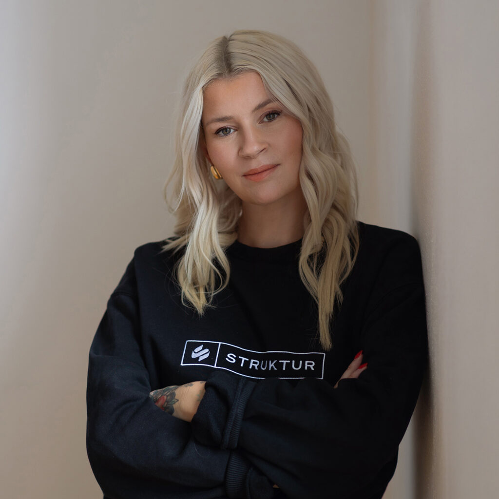 Person mit blondem Haar in schwarzem Pulli, lächelt leicht und steht vor neutralem Hintergrund.