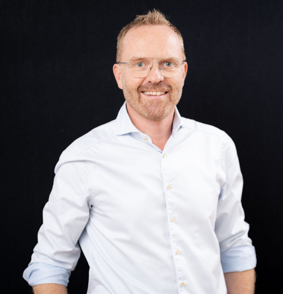 Mann in weißem Hemd vor schwarzem Hintergrund lächelt in die Kamera.