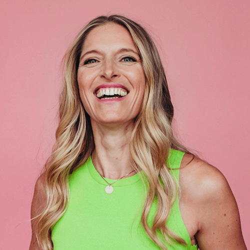 Frau mit langen blonden Haaren lächelt in hellem grünen Oberteil vor rosa Hintergrund.
