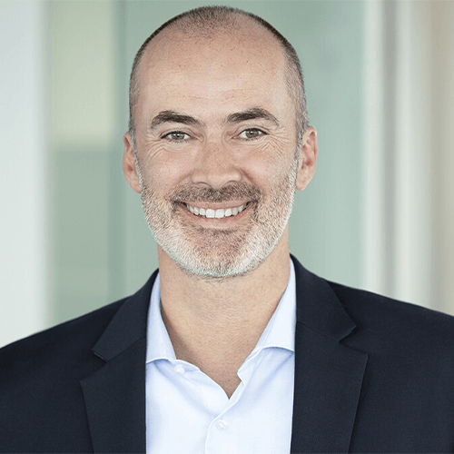 Mann mittleren Alters in Anzug lächelt in die Kamera, steht vor unscharfem Hintergrund in einem modernen Gebäude.