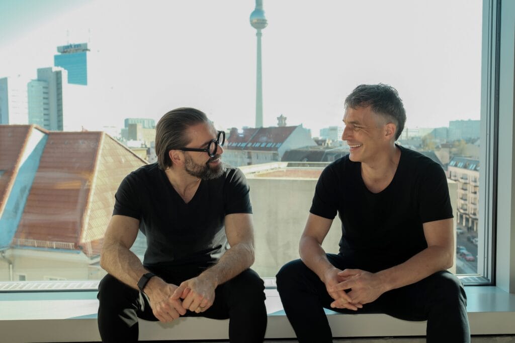 Zwei Männer sitzen lächelnd auf einer Fensterbank vor einer Stadt-Skyline, tragen schwarze T-Shirts.
