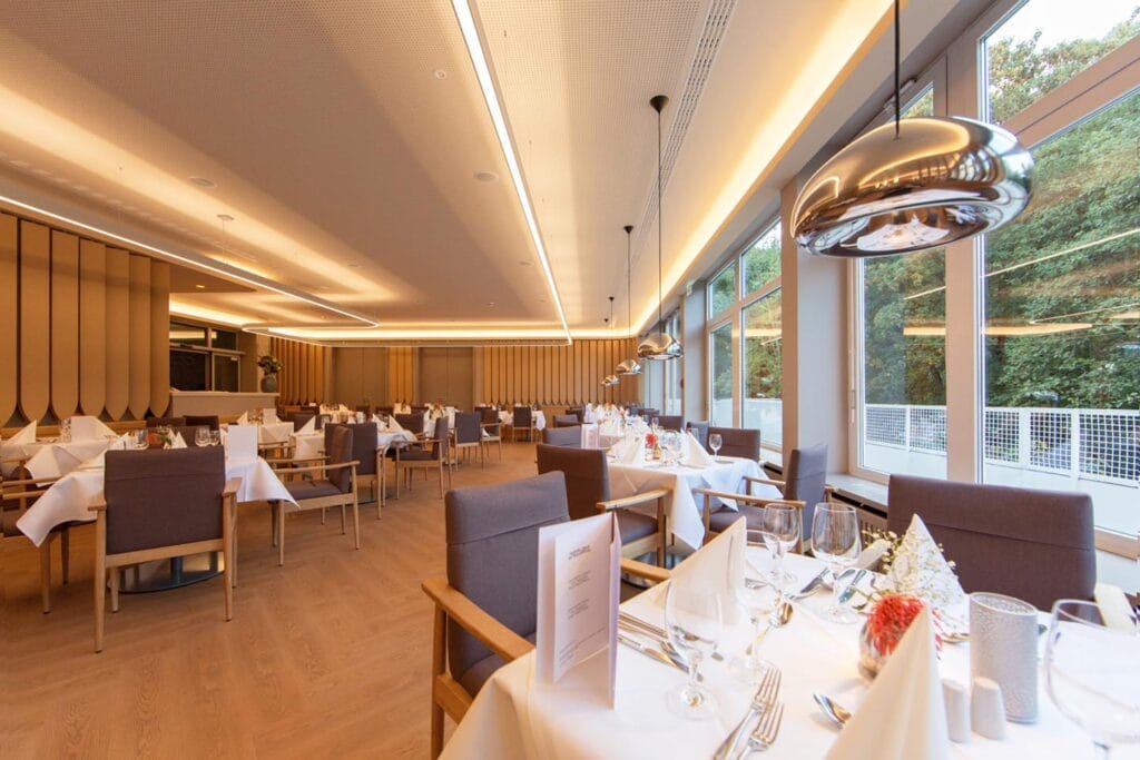 Modernes Restaurant mit hellem Interieur, festlich gedeckten Tischen, weißen Tischdecken, Gläsern und großen Fenstern.