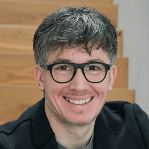 Porträt eines lächelnden Mannes mit schwarzen Brillen, grauen Haaren, vor einer Holztreppe.