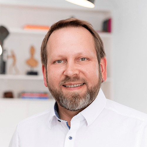 Porträt eines Mannes im weißen Hemd vor Bürohintergrund, freundlich lächelnd.