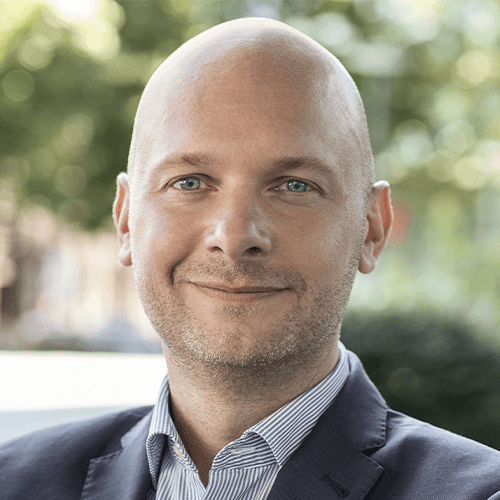 Porträt eines lächelnden Mannes in Business-Outfit im Freien, neutraler Hintergrund.