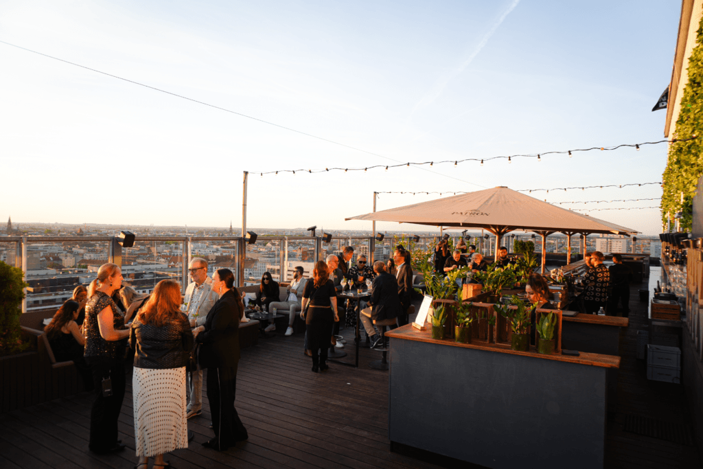 Gruppe von Menschen genießt Rooftop-Party bei Sonnenuntergang mit Stadtblick, Bar und Lichterketten.