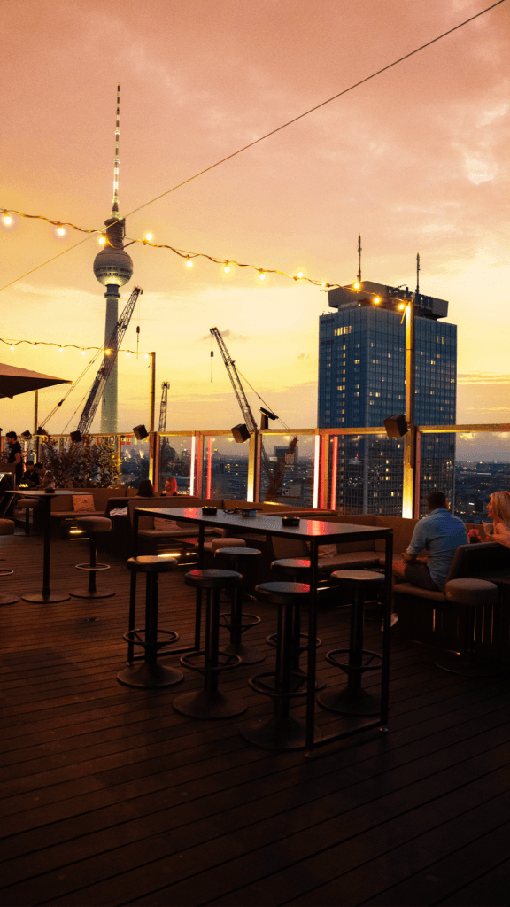 Menschen sitzen auf einer Dachterrasse bei Sonnenuntergang, Blick auf Berliner Skyline mit Fernsehturm und Lichterketten.