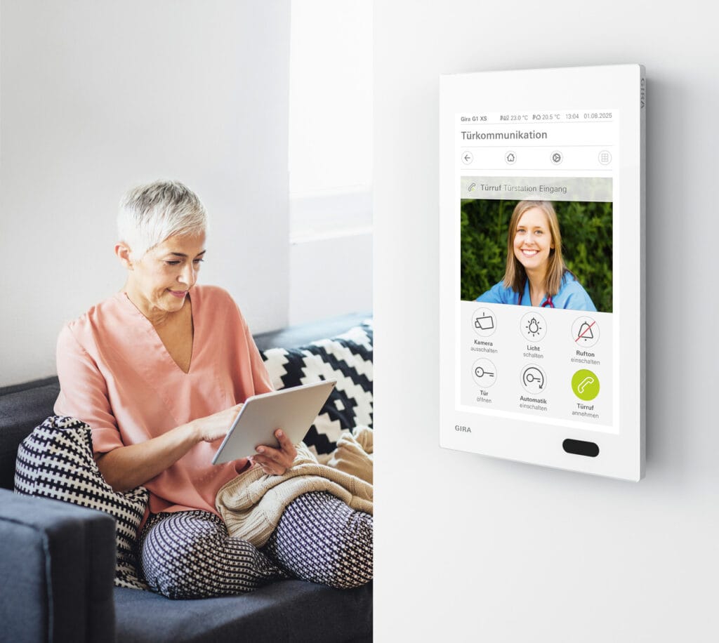Ältere Frau sitzt entspannt auf dem Sofa und nutzt Tablet; Wandpanel zur Türkommunikation mit Display.